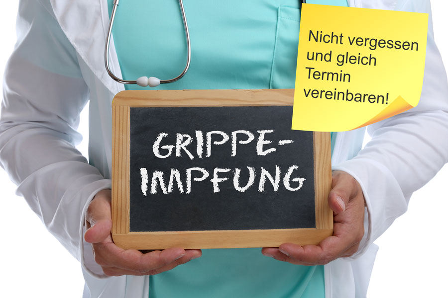 Arzt hält eine Tafel mit der Aufschrift Grippeimpfung und ein aufgeklebter Notizzettel fordert auf, an die Impfung zu denken und einen Termin zu vereinbaren